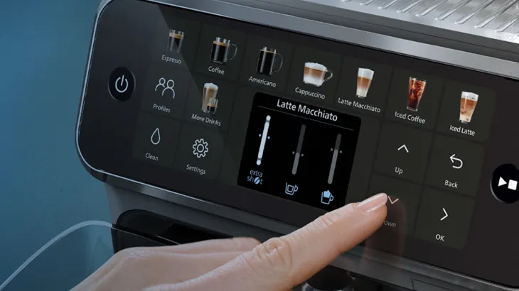 Eine Hand bedient den Touchscreen einer Kaffeemaschine mit Optionen für Espresso, Cappuccino und Latte Macchiato.