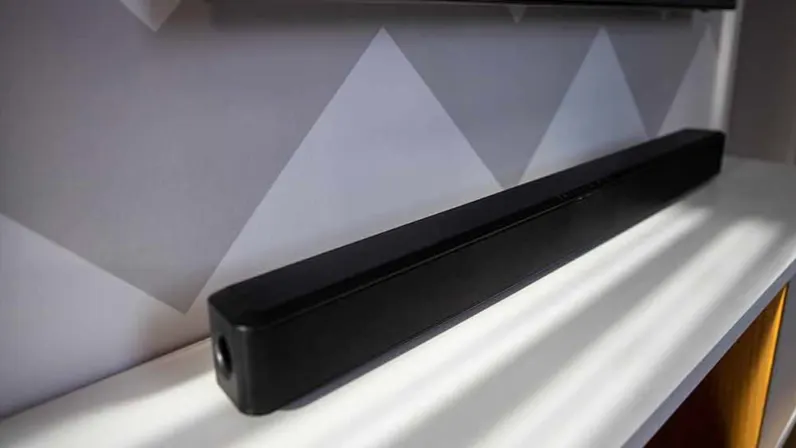 Eine schwarze Soundbar liegt auf einem weißen Regal vor einer Wand mit grauen Dreiecken.