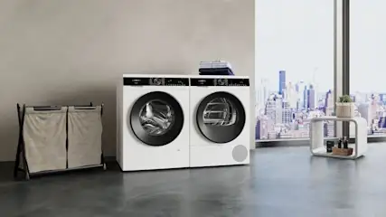 Zwei weiße Waschmaschinen stehen nebeneinander, Wäschekorb links, Tisch mit Pflanze und Stadtblick durch Fenster rechts.