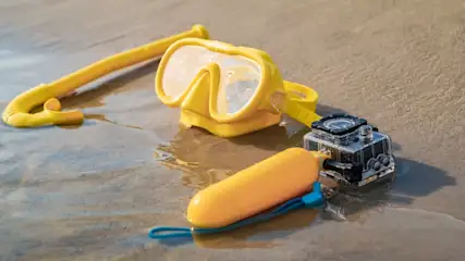 Gelbe Schnorchelausrüstung und Action-Kamera liegen auf Sand. Eine Boje hat ein blaues Band.