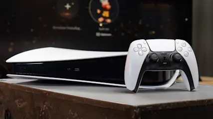 Eine weiße und schwarze Playstation 5 Konsole steht neben einem Controller auf einem braunen Tisch vor einem dunklen Bildschirm.