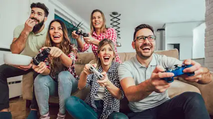 Fünf Personen sitzen auf einem Sofa und spielen Videospiele mit Controllern, während sie lachen.