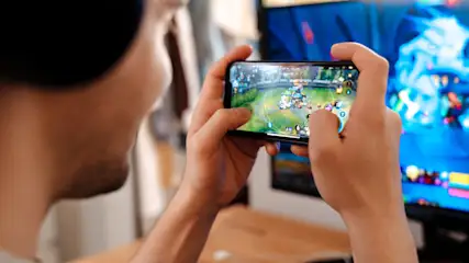 Eine Person mit Kopfhörern hält ein Handy im Querformat, spielt ein Spiel; ein Monitor ist im Hintergrund sichtbar.