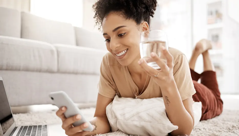 Frau liegt auf Teppich mit Laptop, hält ein Smartphone und ein Glas Wasser in den Händen. Kissen unter dem Oberkörper.