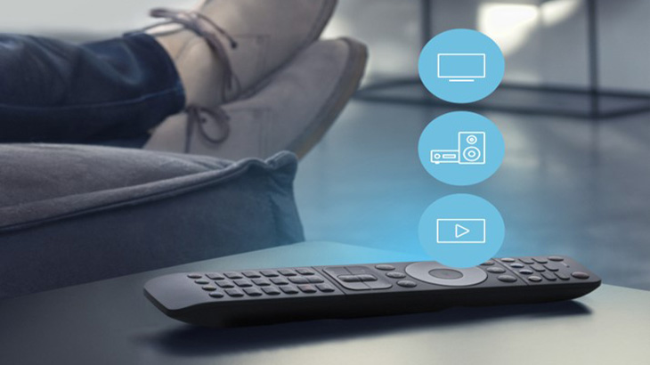 Eine Fernbedienung liegt auf einem Tisch vor einem Sofa, auf dem Füße liegen. Im Hintergrund sind blaue Kreise mit Symbolen.