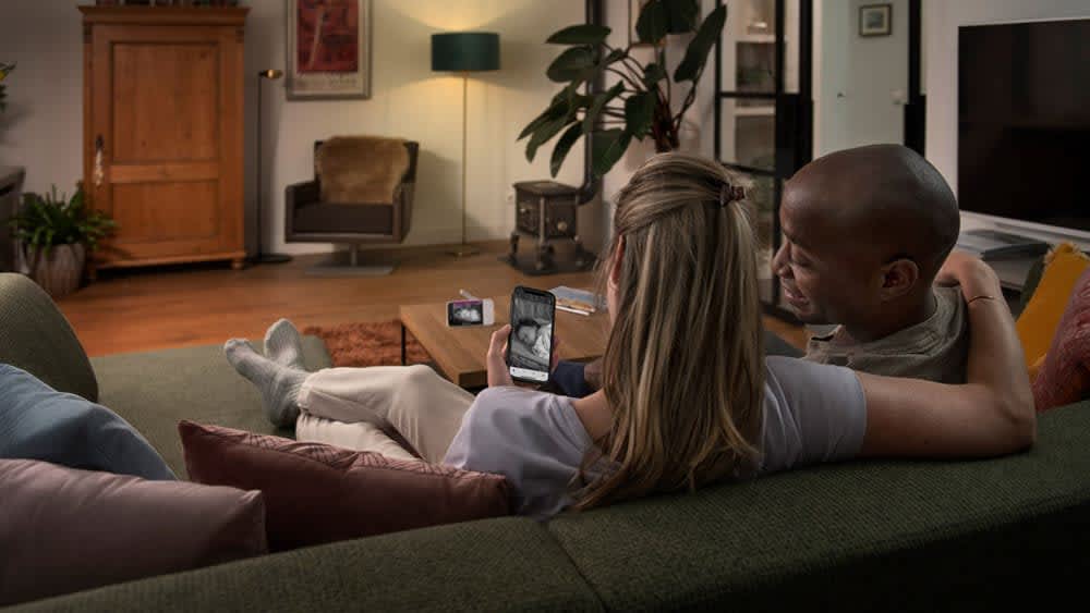 Mann und Frau sitzen auf dem Sofa und beobachten ihr Baby per Smartphone