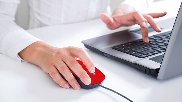 Eine Person tippt auf einem Laptop und bedient eine rote Maus auf einem weißen Tisch.