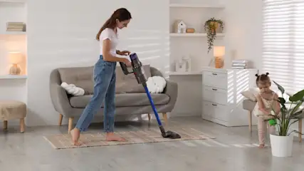Eine Frau staubsaugt einen Teppich mit einem blauen Staubsauger, während ein Kind in der Nähe eine Zimmerpflanze gießt.