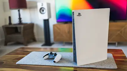 Ein weißes Spielkonsolensystem steht auf einem Holztisch. Ein Controller liegt daneben. Im Hintergrund sind Lautsprecher und ein Fernseher zu sehen.