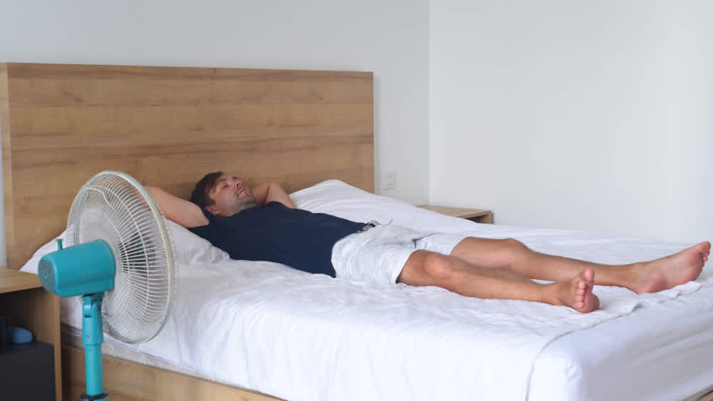 Ein Mann liegt auf dem Bett und ein Ventilator steht daneben.