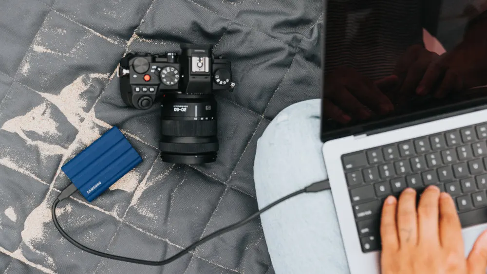 Kamera, blaue Festplatte und Laptop auf einer grauen, sandbedeckten Decke. Eine Person tippt auf dem Laptop.