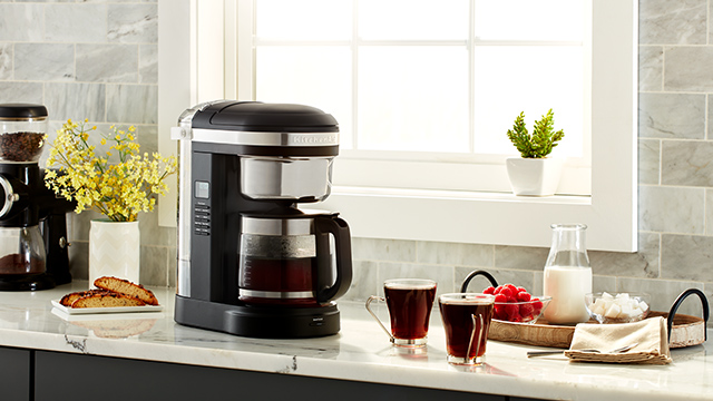 Kaffeemaschine und Mühle auf einer Küchentheke mit Tassen, Milch, Beeren und Gebäck vor einem Fenster.