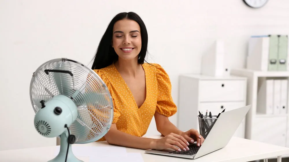 Eine Frau tippt auf einem Laptop an einem weißen Schreibtisch, neben einem blauen Ventilator vor weißem Hintergrund.