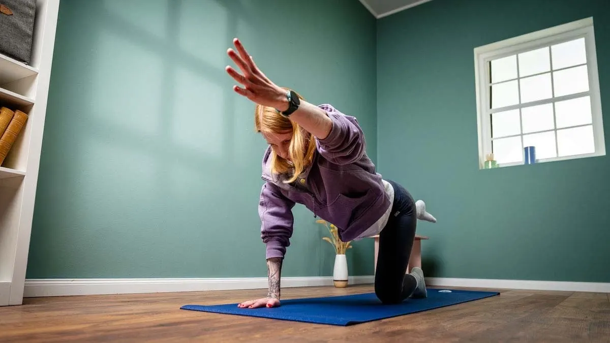 Eine Person auf einer blauen Matte macht Yoga vor einer grünen Wand mit Fenster und einem weißen Regal.