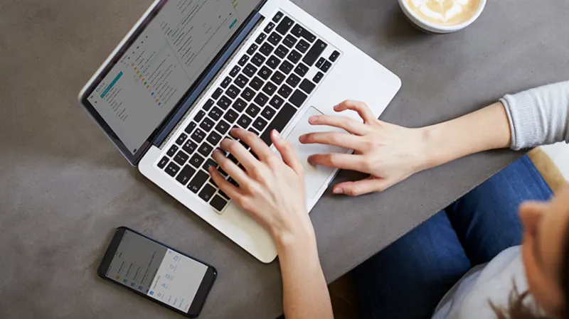 Draufsicht auf Laptop mit tipppenden Händen, Handy, Kaffeetasse auf grauem Tisch. Person trägt Jeans und grauen Pullover.