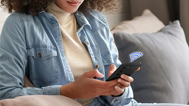 Μια γυναίκα με ένα smartphone στο χέρι της κάθεται σε έναν καναπέ. Πάνω από το smartphone υπάρχει ένα σύμβολο Wi-Fi που υποδεικνύει μια ασύρματη σύνδεση.