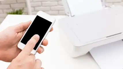 Eine Hand tippt auf ein weißes Smartphone mit schwarzem Bildschirm. Daneben steht ein weißer Drucker auf einem Tisch.