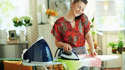 Eine Frau bügelt mit einem Bügeleisen auf einem Bügelbrett und telefoniert. Im Hintergrund sind Blumen zu sehen.