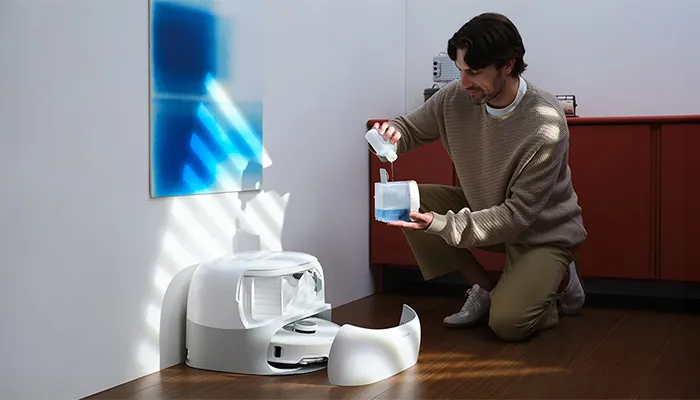 Ein Mann füllt einen Behälter über einer weißen Roboter-Basisstation auf einem Holzboden. An der Wand hängt blaue Kunst.