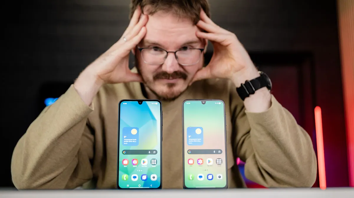 Das Samsung Galaxy A16 (rechts) und das Samsung Galaxy A26 stehen mit eingeschaltetem Display nebeneinander auf dem Tisch. Michael fasst sich im Hintergrund mit den Händen an den Kopf und schaut kritisch.