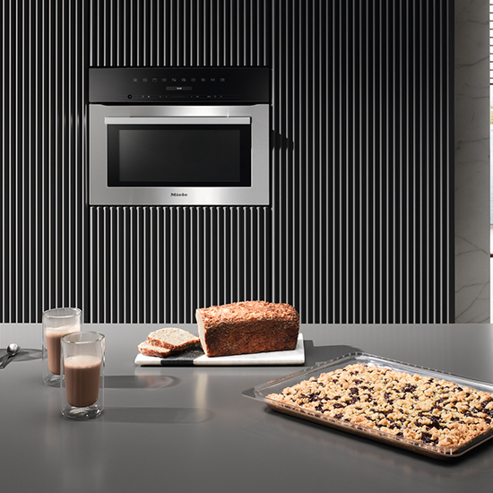 Auf einer Küchentheke stehen Getränke, Brot auf einem Brett und ein Backblech. Ein Ofen ist in eine Wand mit Streifen eingebaut.