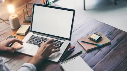 Eine Person tippt auf einem Laptop auf einem Holztisch mit Notizbüchern, Stiften und einer Lampe.