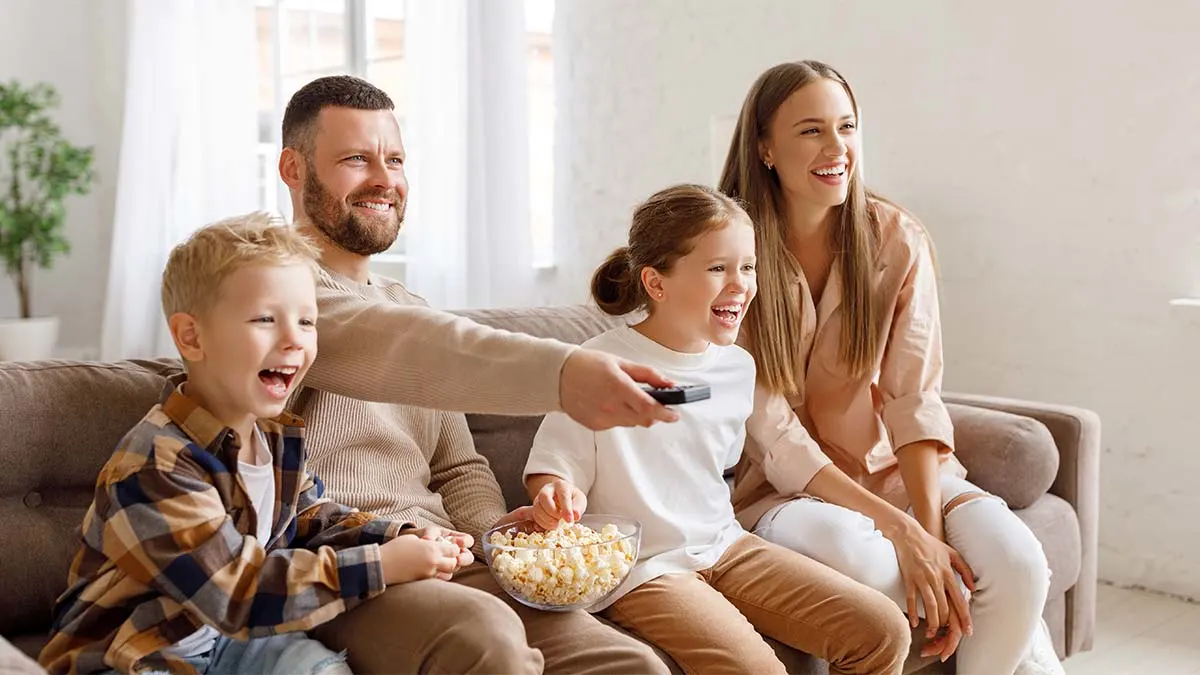 Eine vierköpfige Familie sitzt auf dem Sofa, während der Vater mit der Fernbedienung umschaltet.
