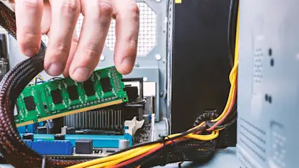 Eine Hand hält einen grünen RAM-Riegel über ein blaues Motherboard in einem offenen Computergehäuse. Kabel sind sichtbar.