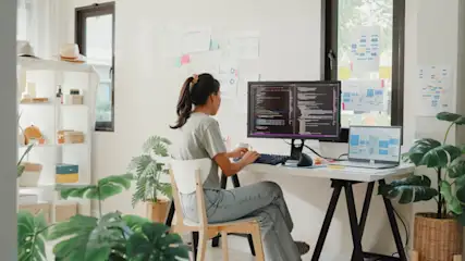Eine Person sitzt an einem Schreibtisch mit Computern, umgeben von Pflanzen und Regalen in einem hellen Raum.