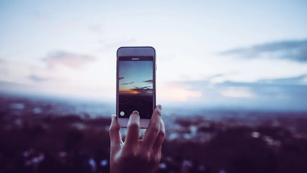 Eine Hand hält ein Smartphone, das einen Sonnenuntergang über einer Stadt auf dem Bildschirm anzeigt. Der Himmel ist hellblau.