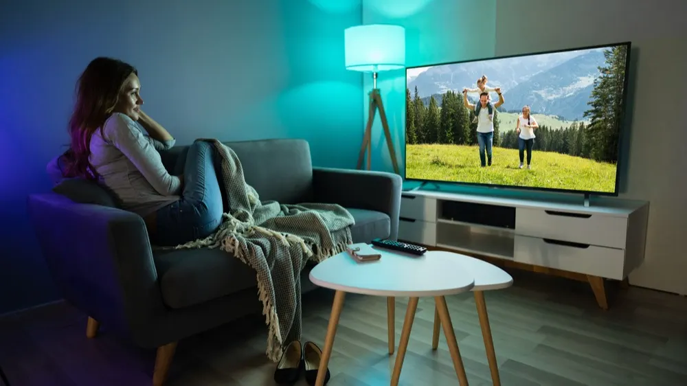 Frau sitzt auf grauem Sofa mit Decke, schaut TV. Tisch, Fernseher und Lampe sind im Raum. Auf dem Bildschirm sind Leute in der Natur.