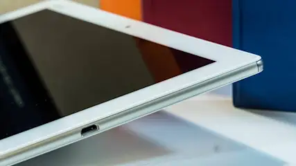 Nahaufnahme eines weißen Tablets mit schwarzem Bildschirm, umgeben von orangefarbenen, roten und blauen Objekten.