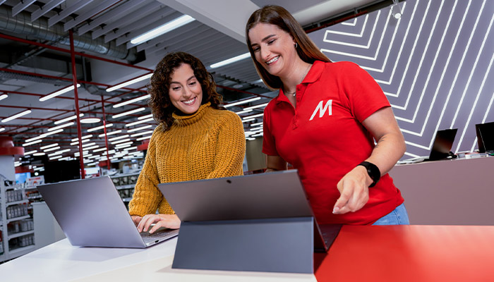 Eine Frau im gelben Pullover bedient einen Laptop neben einer Frau im roten Hemd mit weißem "M" in einem Geschäft.
