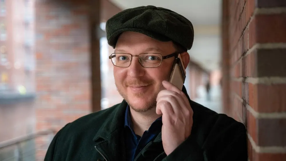 Ein Mann mit Brille und Schirmmütze telefoniert vor einer Ziegelwand. Er trägt ein dunkles Jackett über einem blauen Hemd.