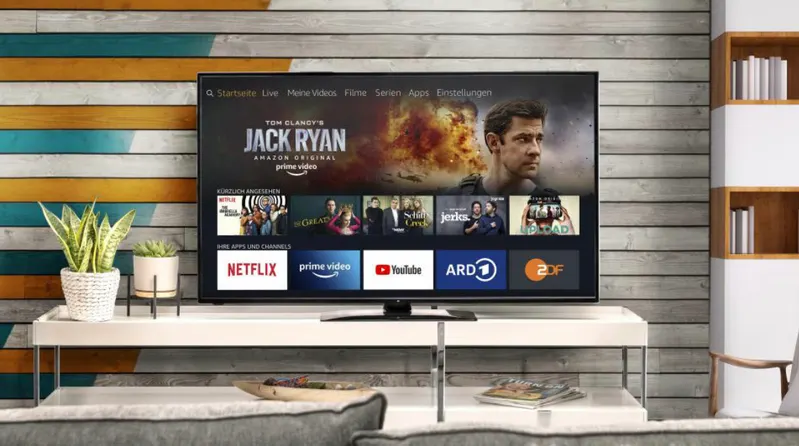 Ein Fernseher zeigt das Menü mit Optionen wie Netflix, Prime Video und YouTube. Pflanzen stehen auf einem weißen Tisch vor einer Wand.