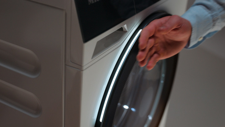 Eine Hand in einem hellblauen Hemd öffnet die Tür einer weißen Waschmaschine mit schwarzem Rand.