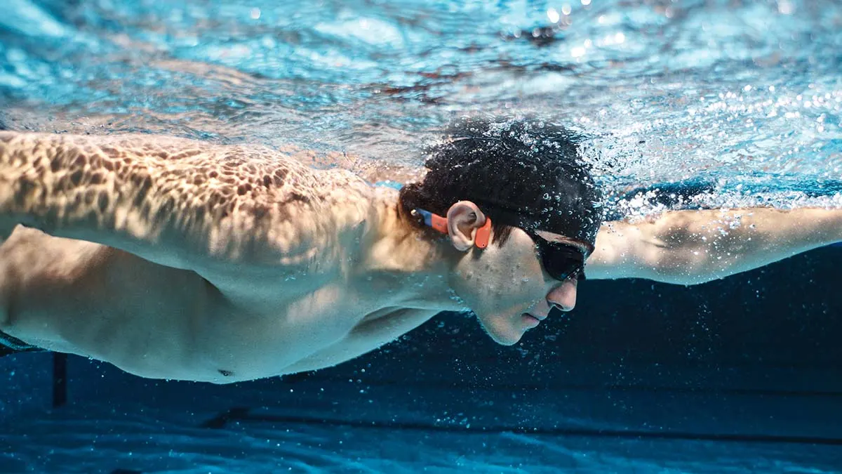 Ein Schwimmer mit Brille und orangen Ohrstöpseln unter Wasser, Arme ausgestreckt, in einem Schwimmbecken.