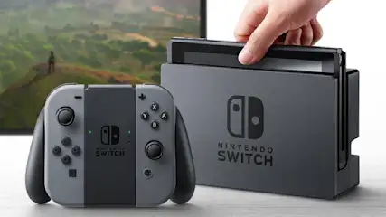 Eine graue Nintendo Switch mit Controllern und Dock. Eine Hand legt die Konsole in das Dock. Ein Bildschirm zeigt eine grüne Landschaft.