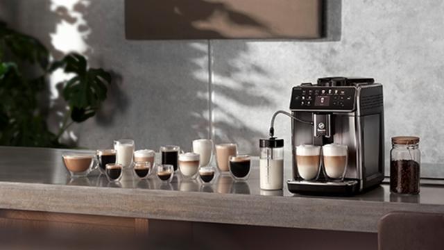 Kaffeemaschine neben Gläsern mit Kaffee, Milch und Bohnen auf einer Küchentheke vor einer Wand und einer Pflanze.