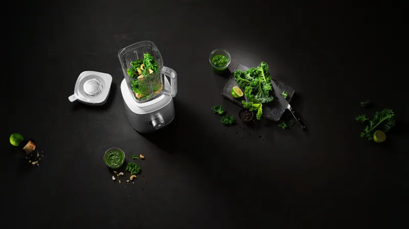 Ein Mixer mit Grünzeug, ein Glas grüner Flüssigkeit, Kohlblätter, Limetten und eine Gabel liegen auf einer schwarzen Oberfläche.