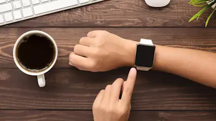 Dunkler Kaffee, Tastatur, Maus, Pflanze und eine Hand mit Smartwatch auf einem braunen Holztisch.