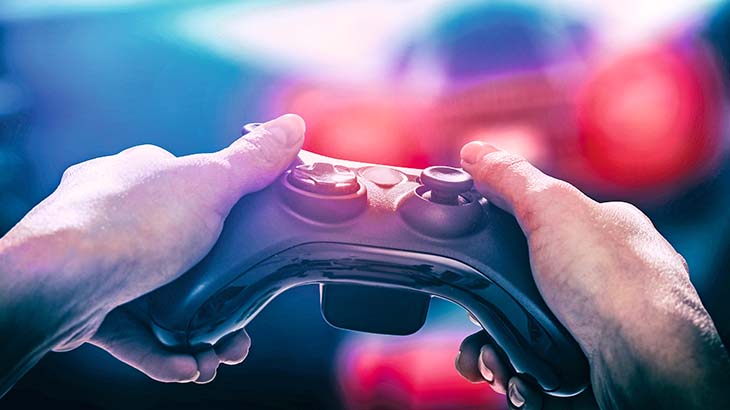 Zwei Hände halten einen schwarzen Controller vor einem verschwommenen Hintergrund mit blauen und roten Farbtönen. Die Hände scheinen das Gerät zu steuern.