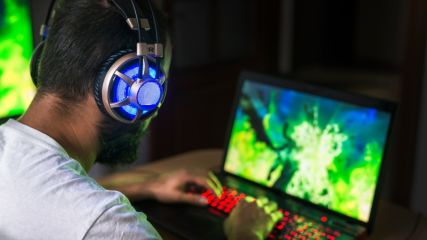 Junger Mann sitzt mit einem blau leuchtenden Headset an einem Laptop und spielt ein Game. 