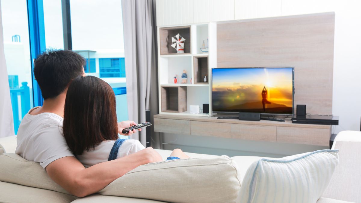Ein Paar sitzt auf einem Sofa und schaut fern. Der Fernseher zeigt ein sonniges Strandbild. Ein Regal steht daneben.