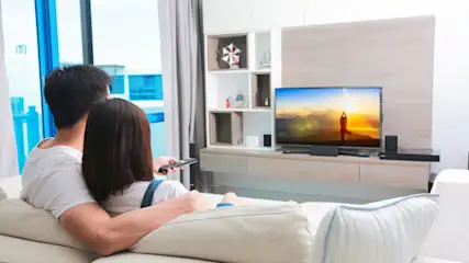 Ein Paar sitzt auf einem Sofa und schaut fern. Der Fernseher zeigt ein sonniges Strandbild. Ein Regal steht daneben.