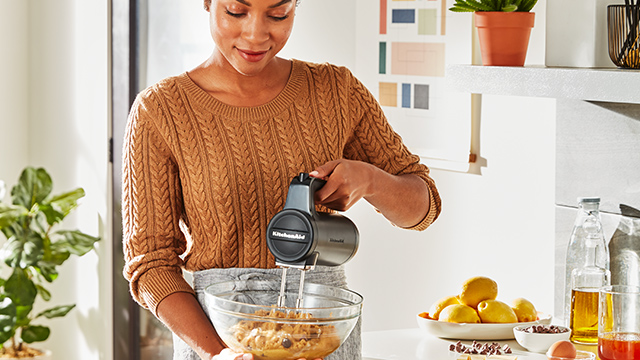 Eine Frau mischt Teig in einer Glasschüssel mit einem grauen Handmixer in einer hellen Küche.