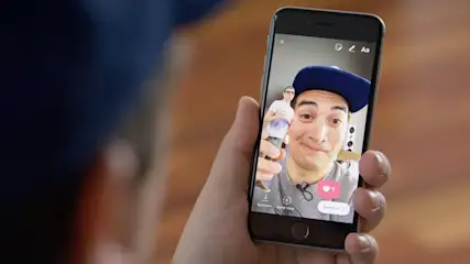 Eine Hand hält ein Smartphone, auf dessen Bildschirm ein Mann mit Kappe und eine weitere Person zu sehen sind.
