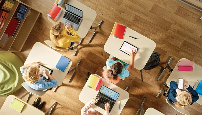 Blick von oben in eine Grundschulklasse. Schüler sitzen an separaten Schreibtischen, vor ihnen liegt jeweils ein Notebook oder ein Tablet.