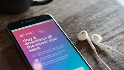 Ein Smartphone mit Apple Music App liegt neben weißen Kopfhörern auf einem Holztisch. Eine schwarze Tasse ist im Hintergrund.