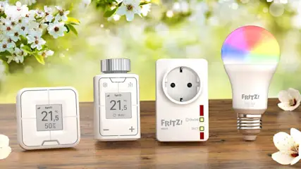 Vier weiße FRITZ!-Smart-Home-Geräte, darunter Thermostate, eine Steckdose und eine Lampe, stehen auf einem Holztisch vor Blumen.